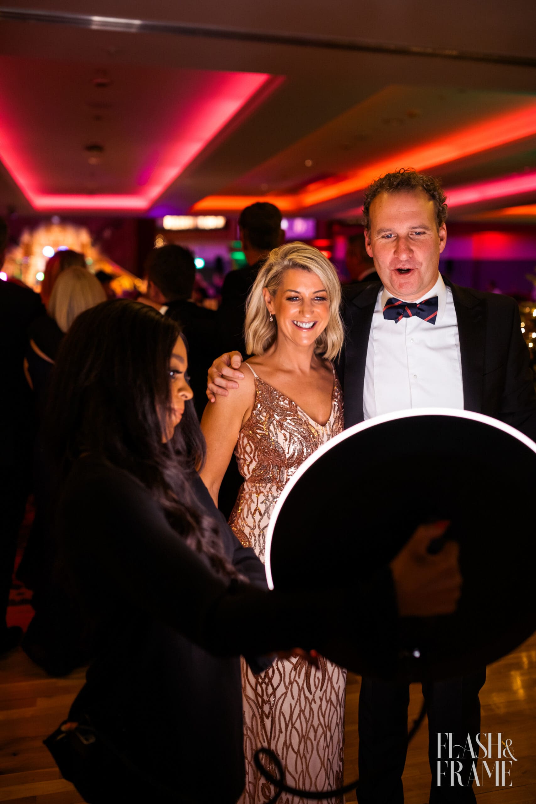 A Flash and Frame booth babe holding a roaming photobooth , a dslr phot station that is on the go through the room, with a group of smiling happy people dressed in gala dress and tux, looking at the screen at their fantastic photo capture.
